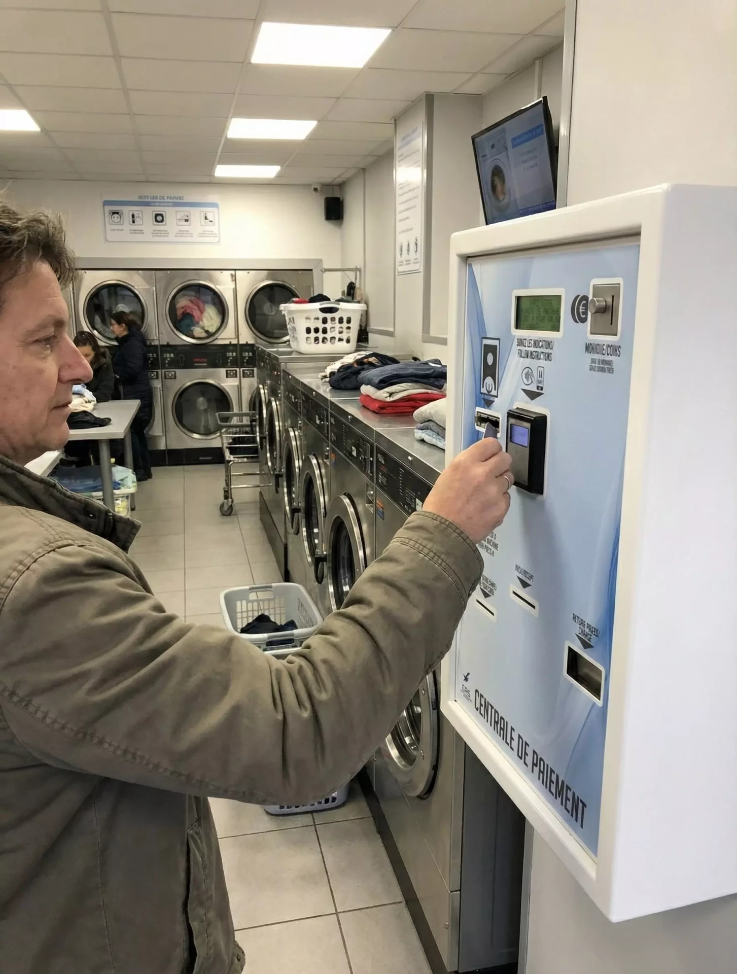 Un homme utilise une machine à laver dans une laverie automatique. Il insère des pièces dans un distributeur. D'autres mac