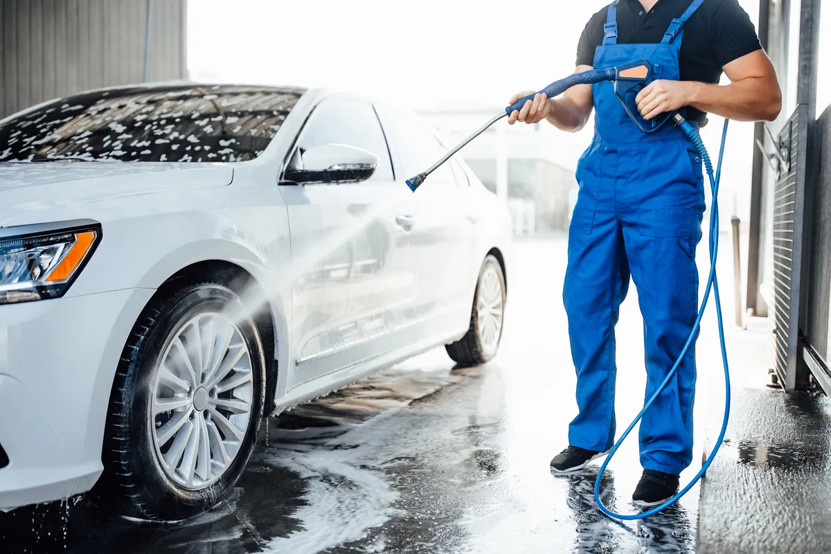 Un technicien en bleu de travail nettoie une voiture avec un tuyau haute pression.